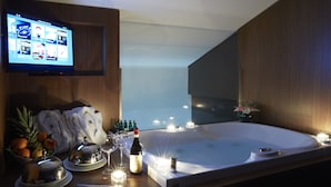 Una habitación moderna con una bañera grande, un televisor montado en la pared y una mesa con toallas, fruta y botellas.