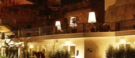 A hotel courtyard at night with a stone wall, potted plants, and a balcony with lights.