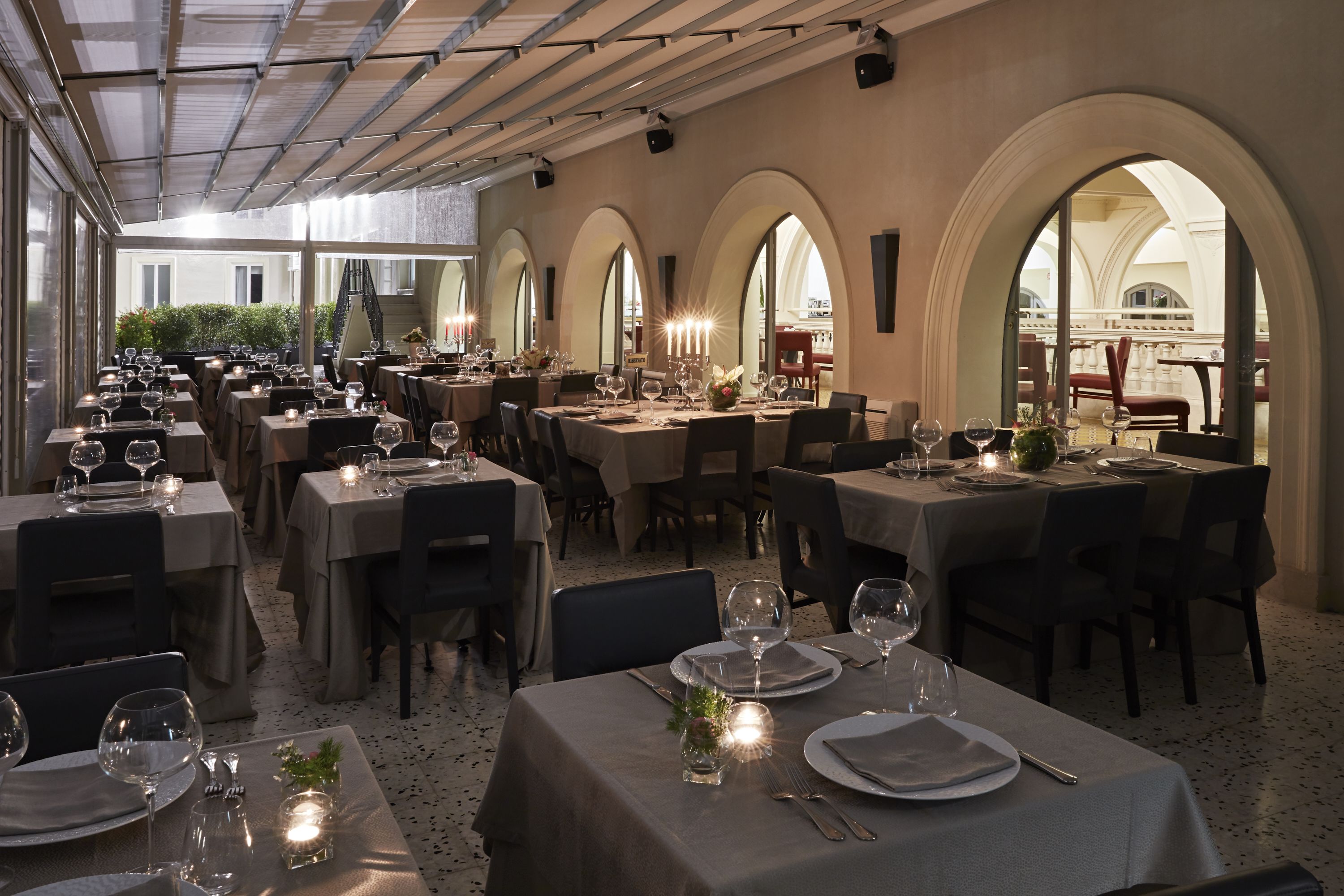 A restaurant with multiple tables set for dining, featuring a covered outdoor area with arched windows and a view of the cityscape.