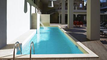 A swimming pool with a wooden deck, metal railings, and a view of a building with a balcony and outdoor seating.