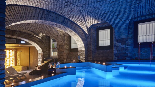 Una piscina interior moderna con techo abovedado de ladrillo, zona de estar con sofá y mesa, y pared de piedra.
