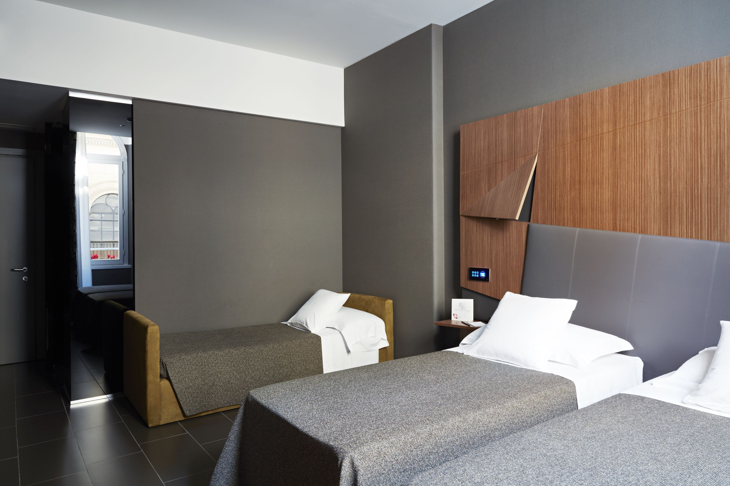 Habitación de hotel con dos camas, cabecero de madera, mesita de noche y vista a un edificio desde una puerta abierta.