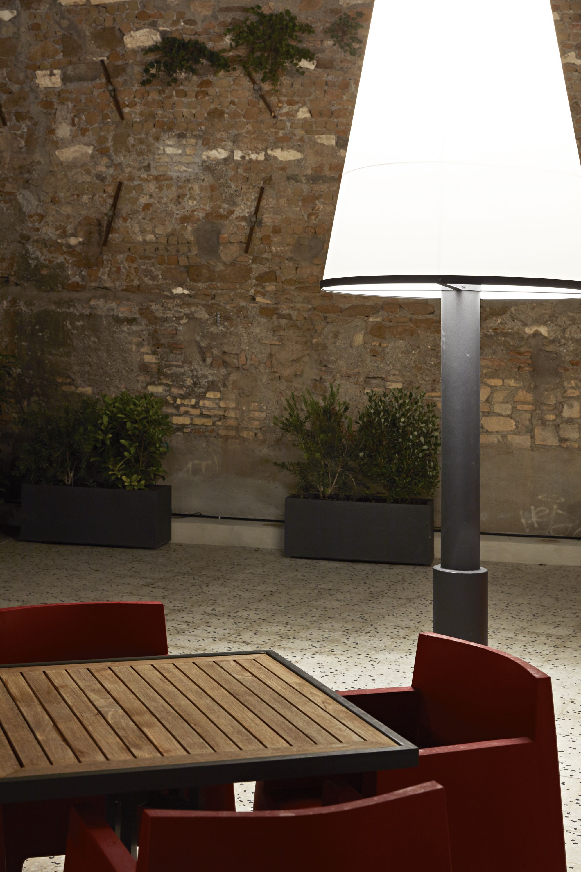 Un espace extérieur aménagé avec une table en bois et des chaises rouges, un lampadaire en métal et des plantes en pot, le tout adossé à un mur de briques.