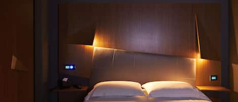 Una habitación de hotel moderna con una cama grande, cabecera de madera, mesitas de noche e iluminación empotrada.