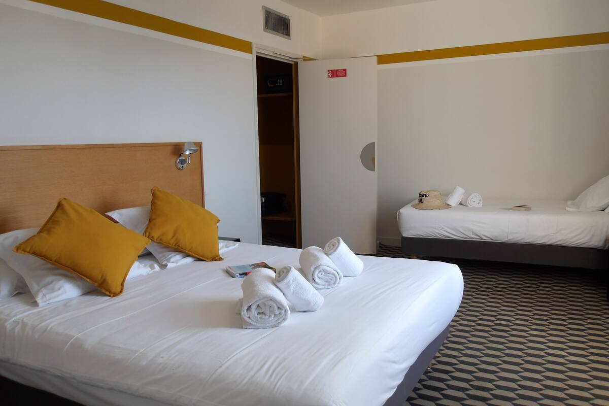 A hotel room with two beds, one with yellow pillows and the other with white pillows. There are towels neatly folded on the beds.
