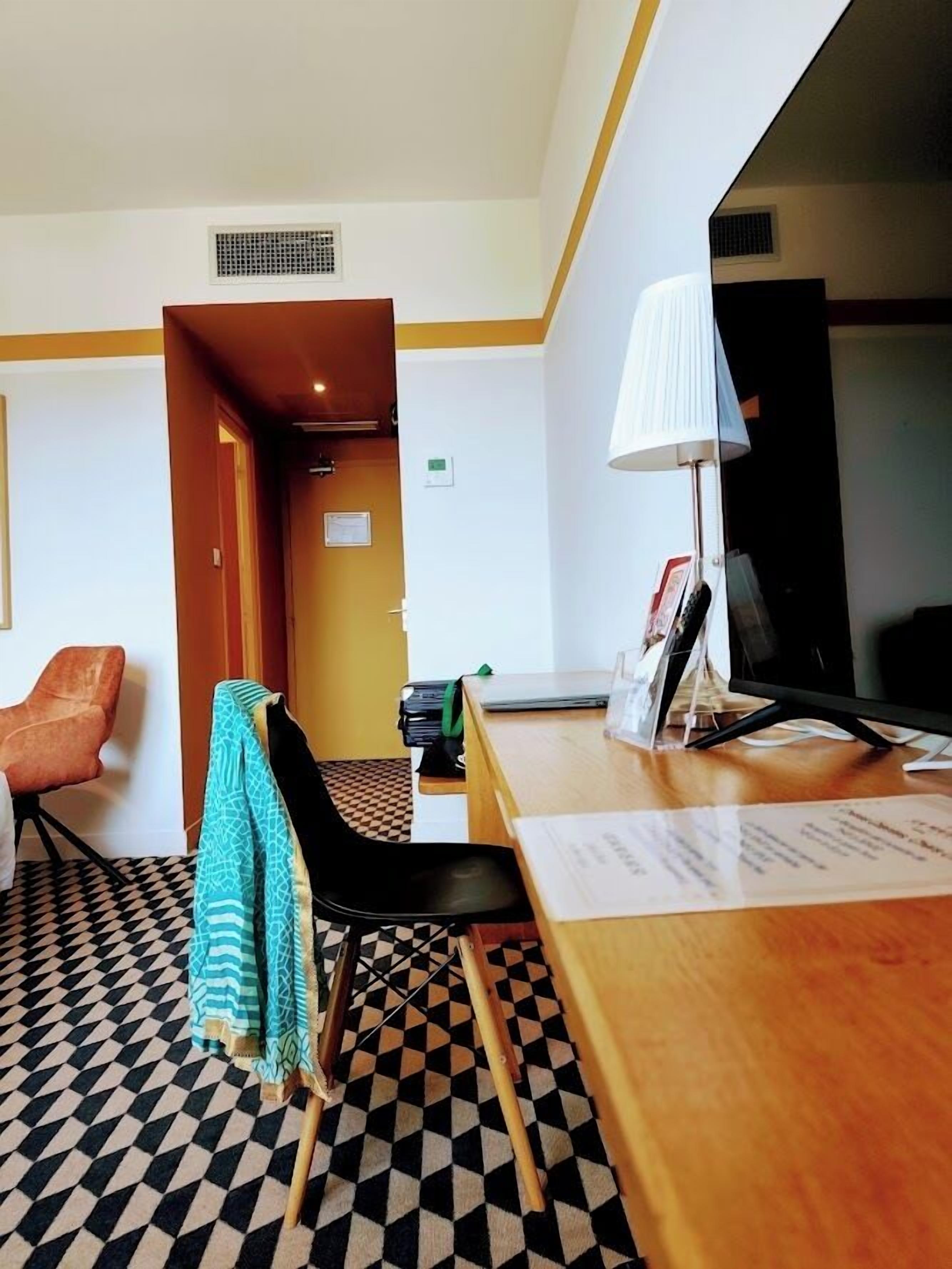 Una camera d'hotel con una reception, una scrivania con una lampada, un televisore, un pavimento decorato e una porta che dà su un'altra stanza.