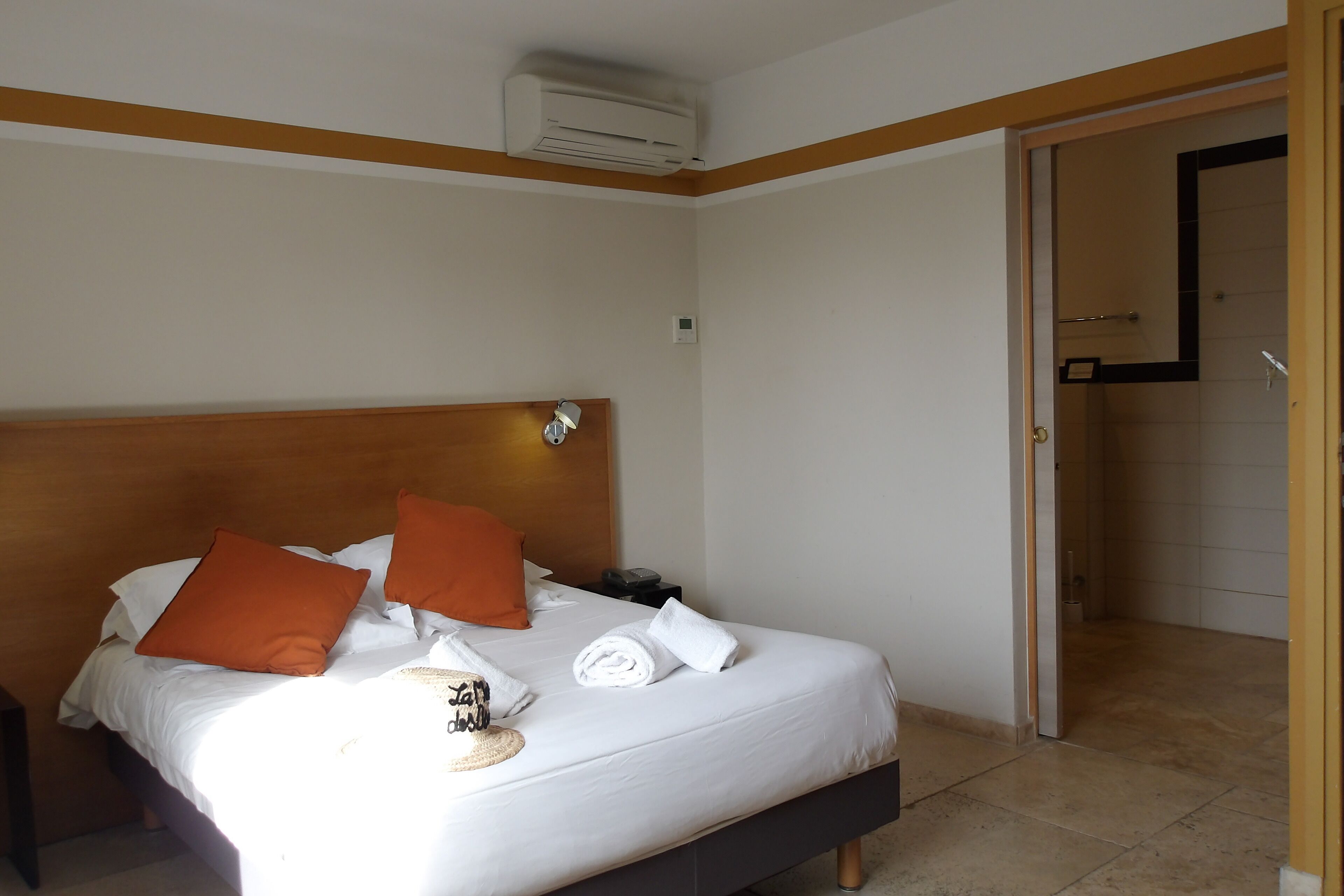 A hotel room with a bed, pillows, a bedside table, an air conditioning unit, and a door leading to a bathroom.