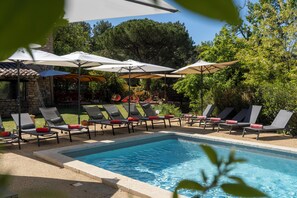 A poolside area with lounge chairs and umbrellas, surrounded by greenery and a stone building. - La Maison des Ocres (Roussillon)