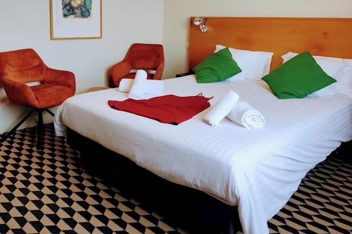 A hotel room with a bed, two chairs, a painting on the wall, and a patterned floor.