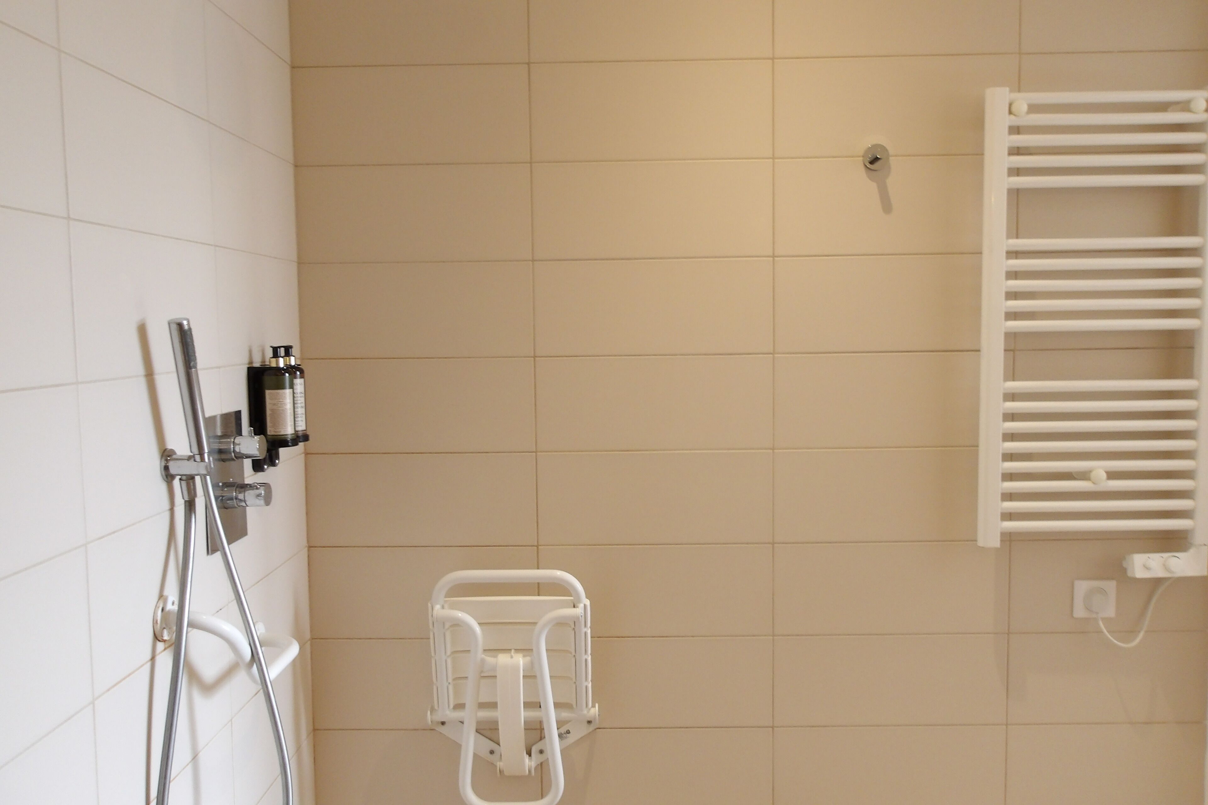 A bathroom with white tiles, a shower with a handheld showerhead, a white towel rack, and a white towel warmer.
