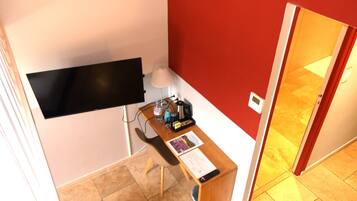 A small room with a desk, a chair, a flat-screen TV mounted on the wall, a lamp, and a door leading to another room.