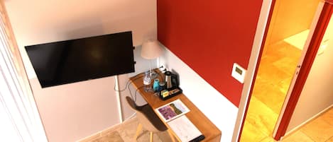 A small room with a desk, a chair, a flat-screen TV mounted on the wall, a lamp, and a door leading to another room.