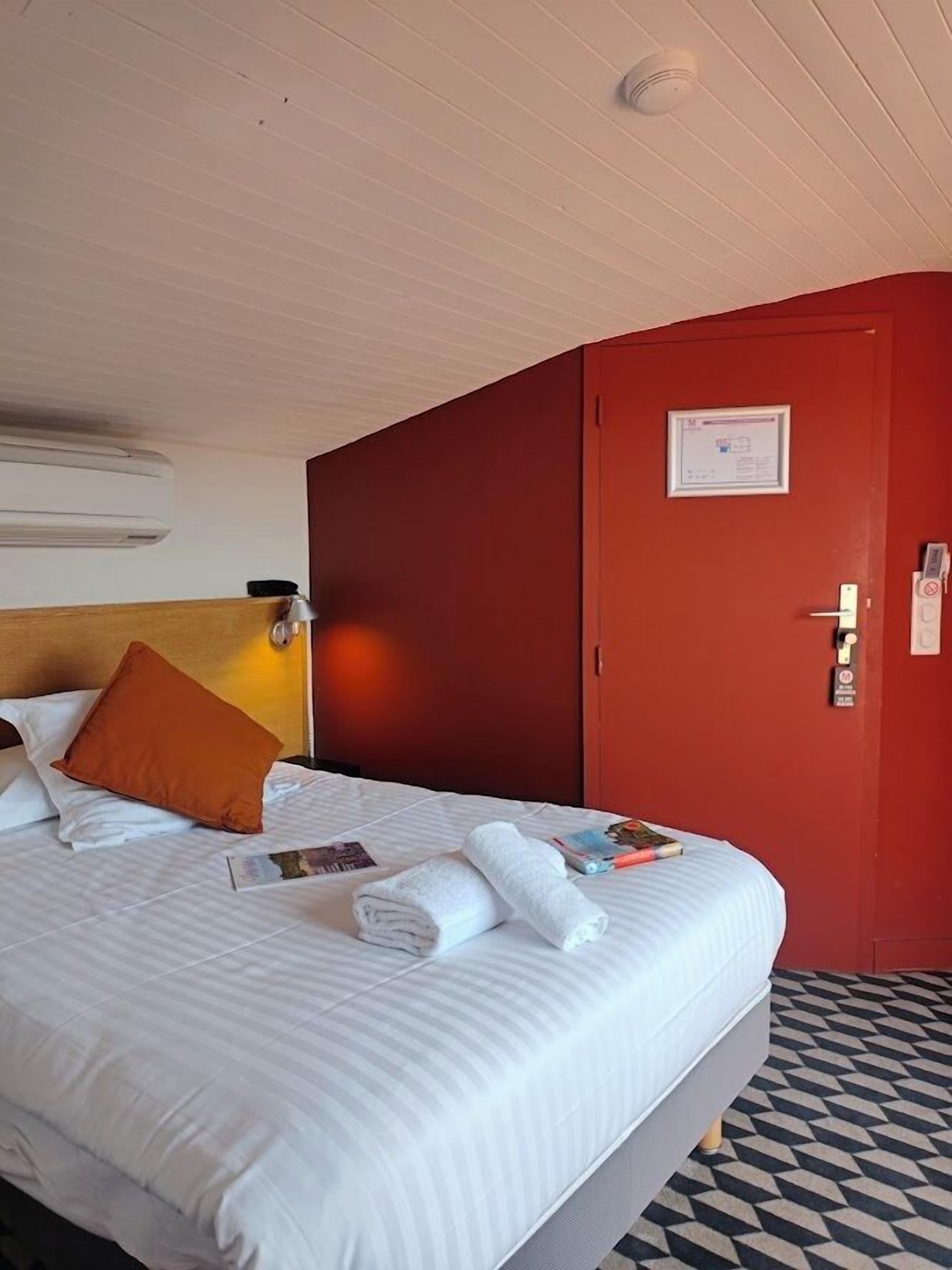Une chambre d’hôtel avec un lit, un mur rouge, une porte avec une pancarte et un sol à motifs.