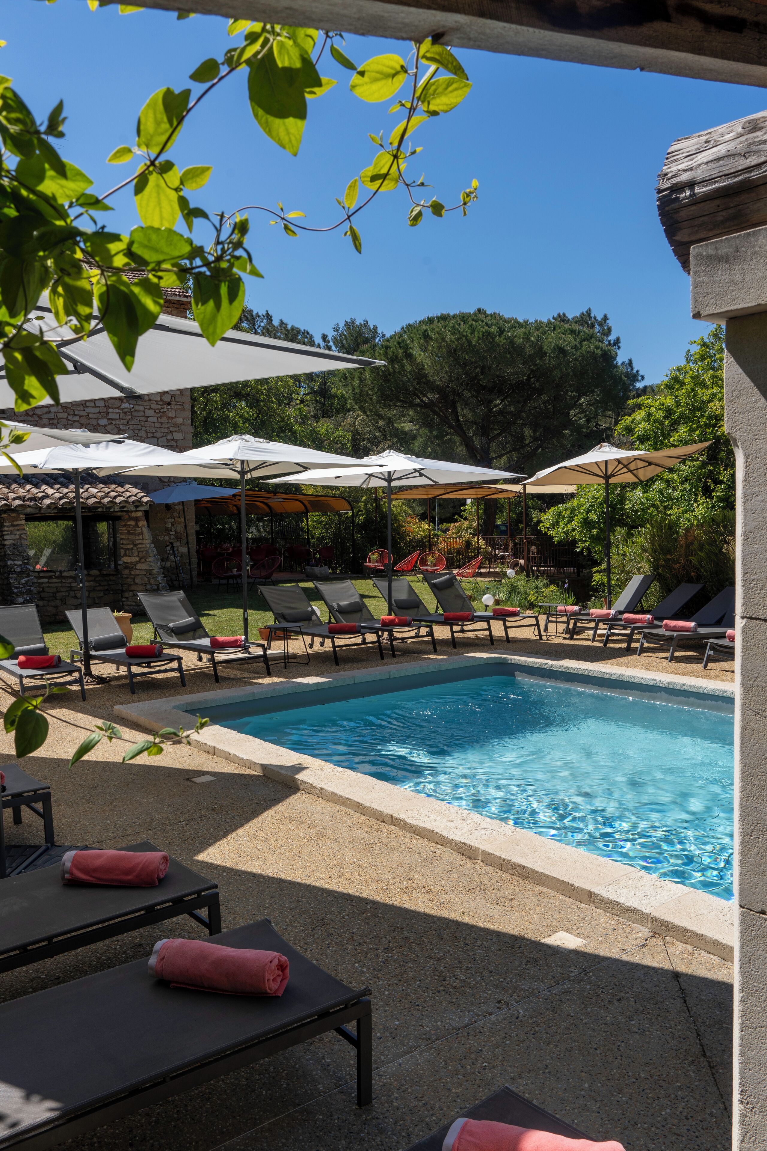 Un espace piscine agrémenté de chaises longues et de parasols, entouré de verdure et bordé d’un bâtiment en pierre.