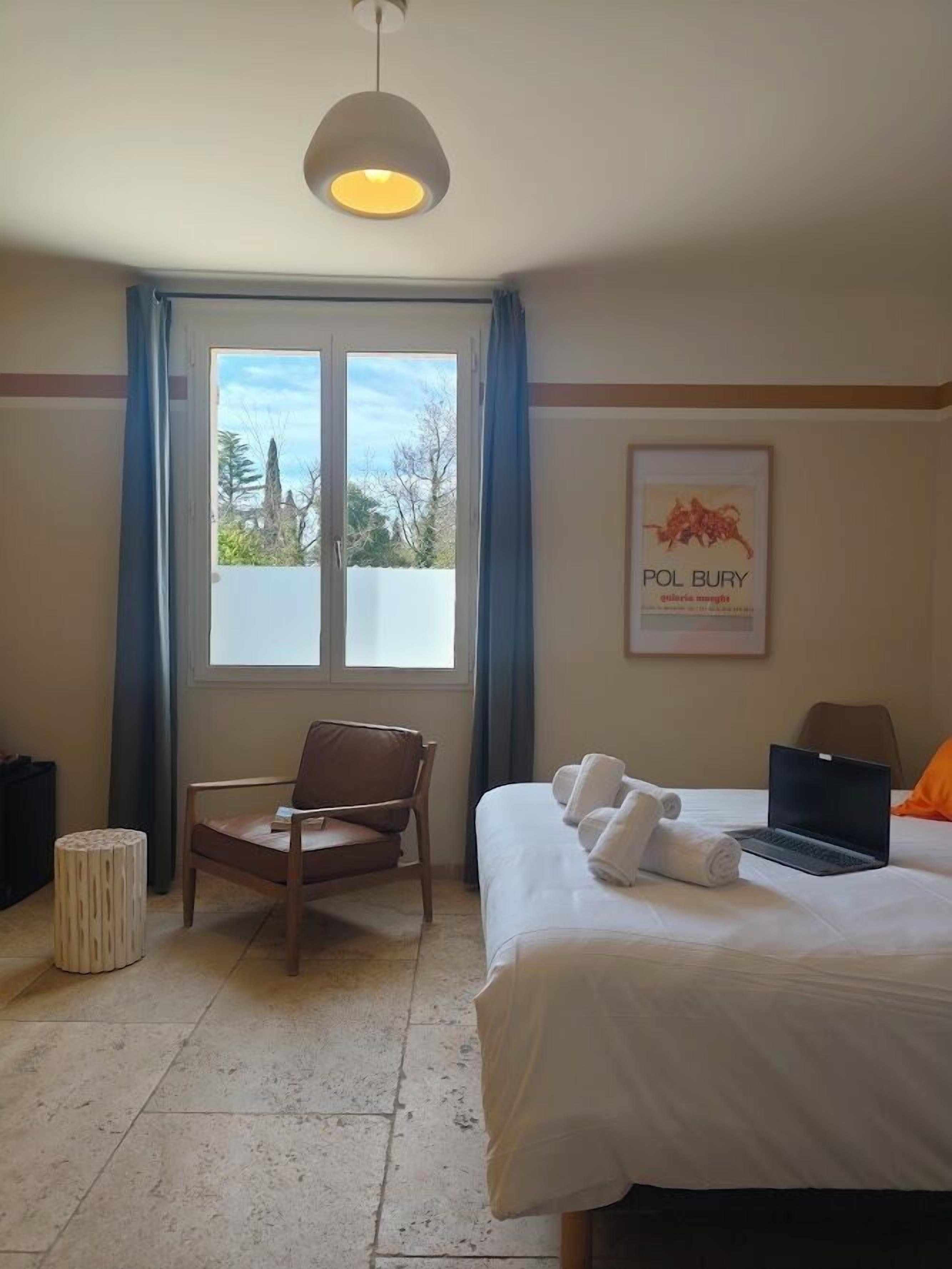 Habitación de hotel con una cama, una silla, una computadora portátil, un cuadro en la pared y un ventanal con cortinas.