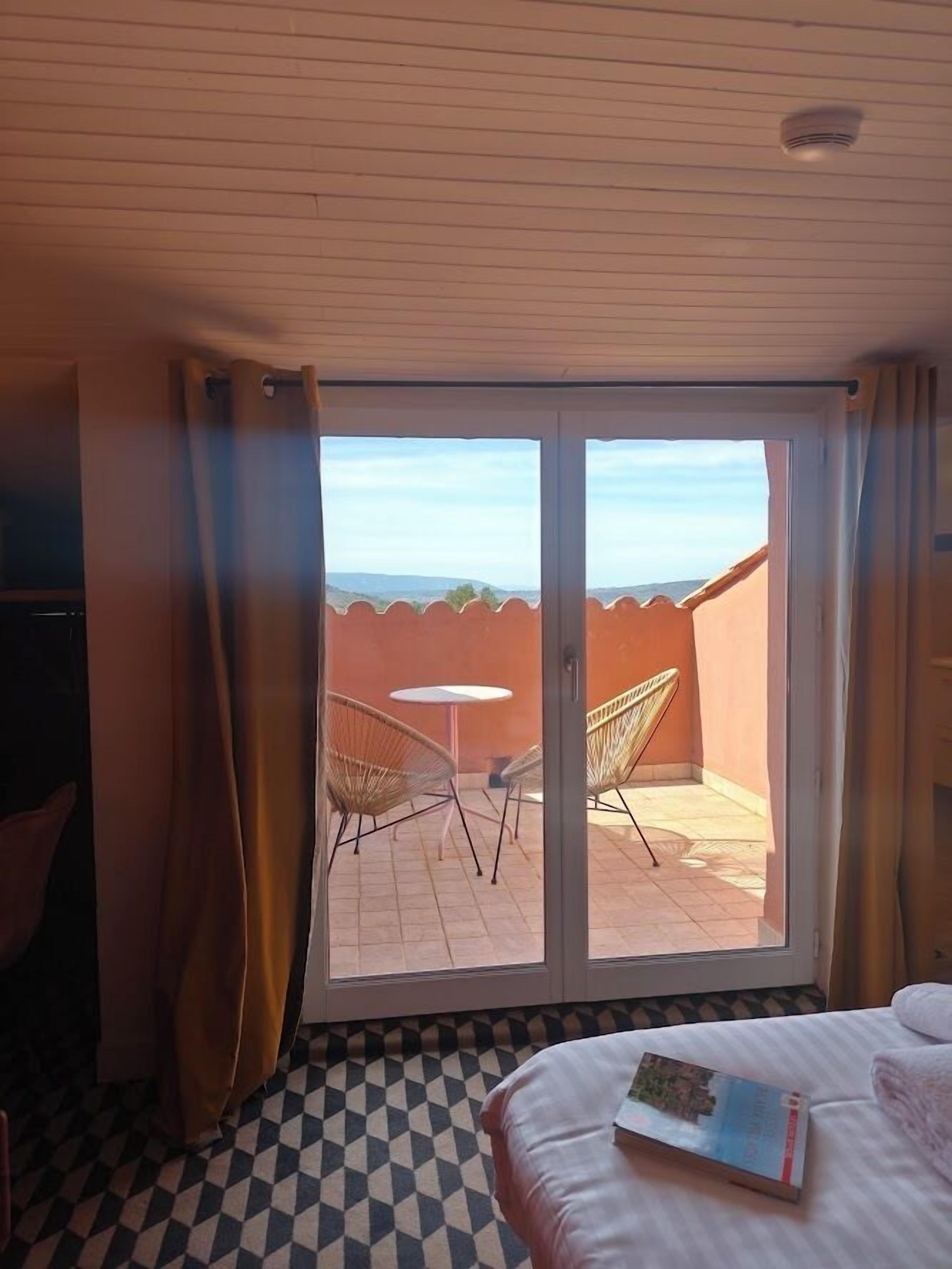 A hotel room with a checkered floor, a bed with a book, a small table, two wicker chairs, and a view of the outdoors through a sliding door.