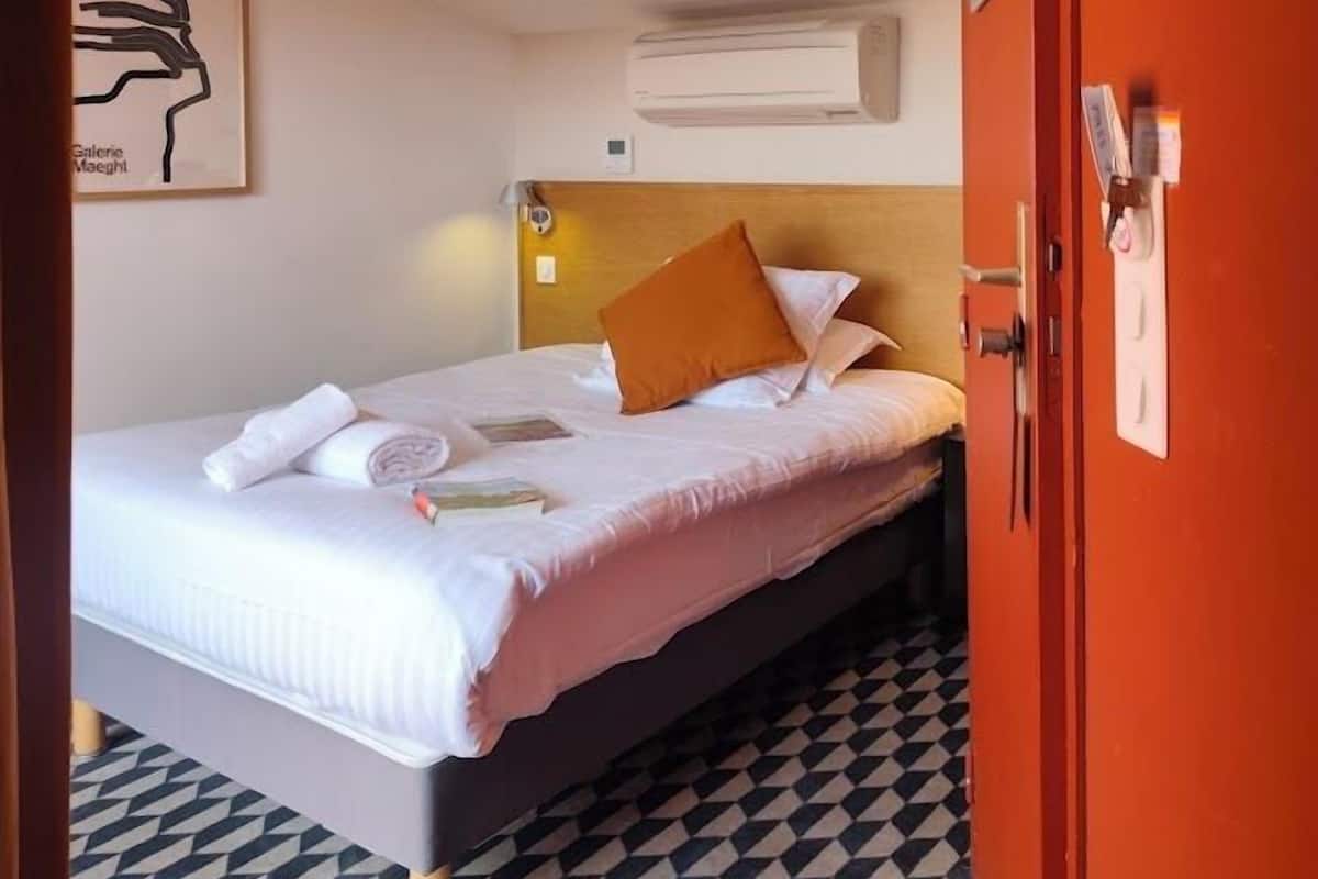 A hotel room with a bed, a headboard, a wall-mounted air conditioner, a framed artwork, and a patterned floor.