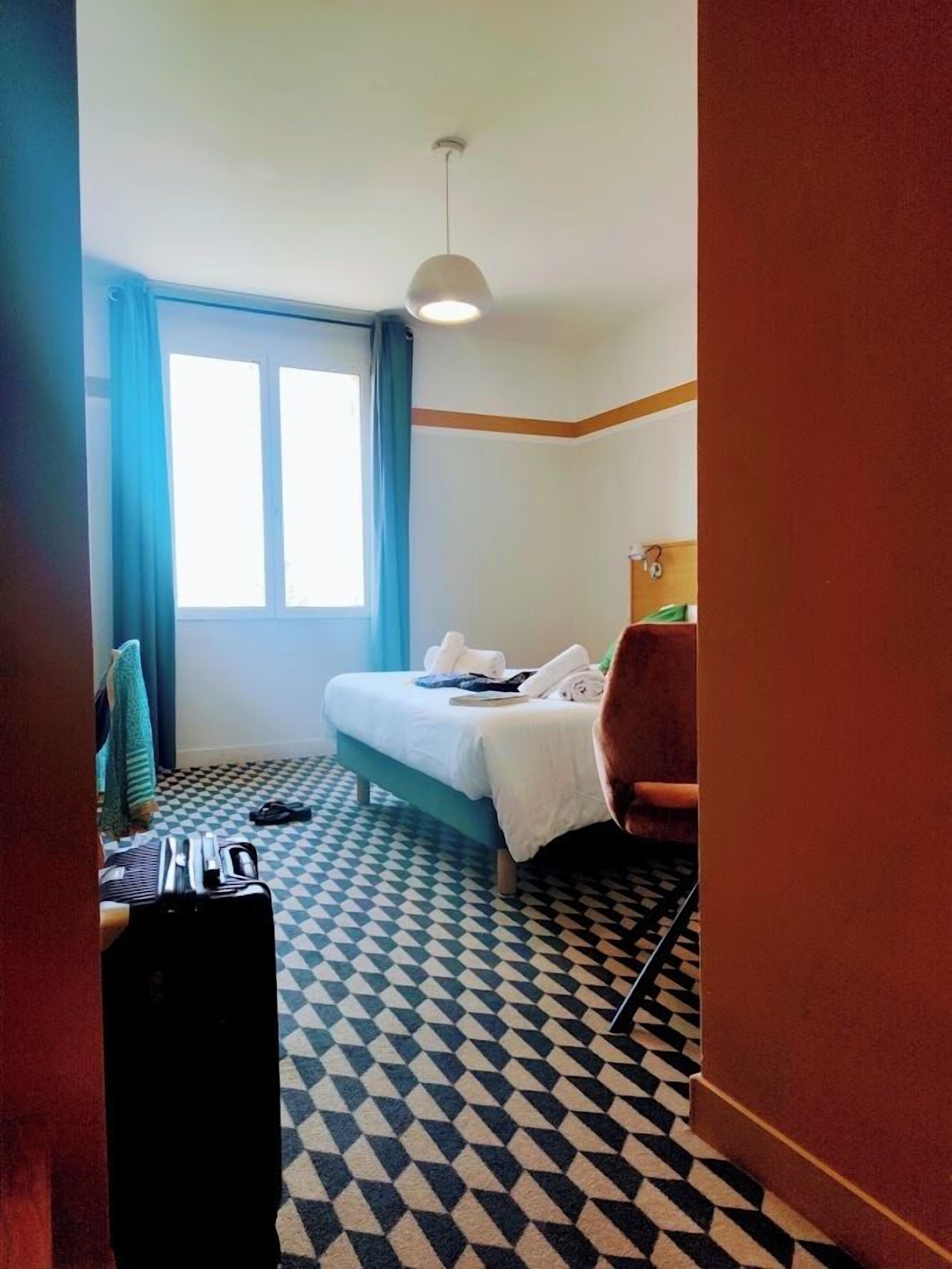 A hotel room with a checkered floor, a bed with white linens, a desk with a chair, and a window with blue curtains.