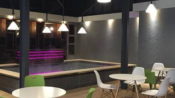 Espaço interior moderno com piscina, cadeiras verdes e brancas, e iluminação pendente.
