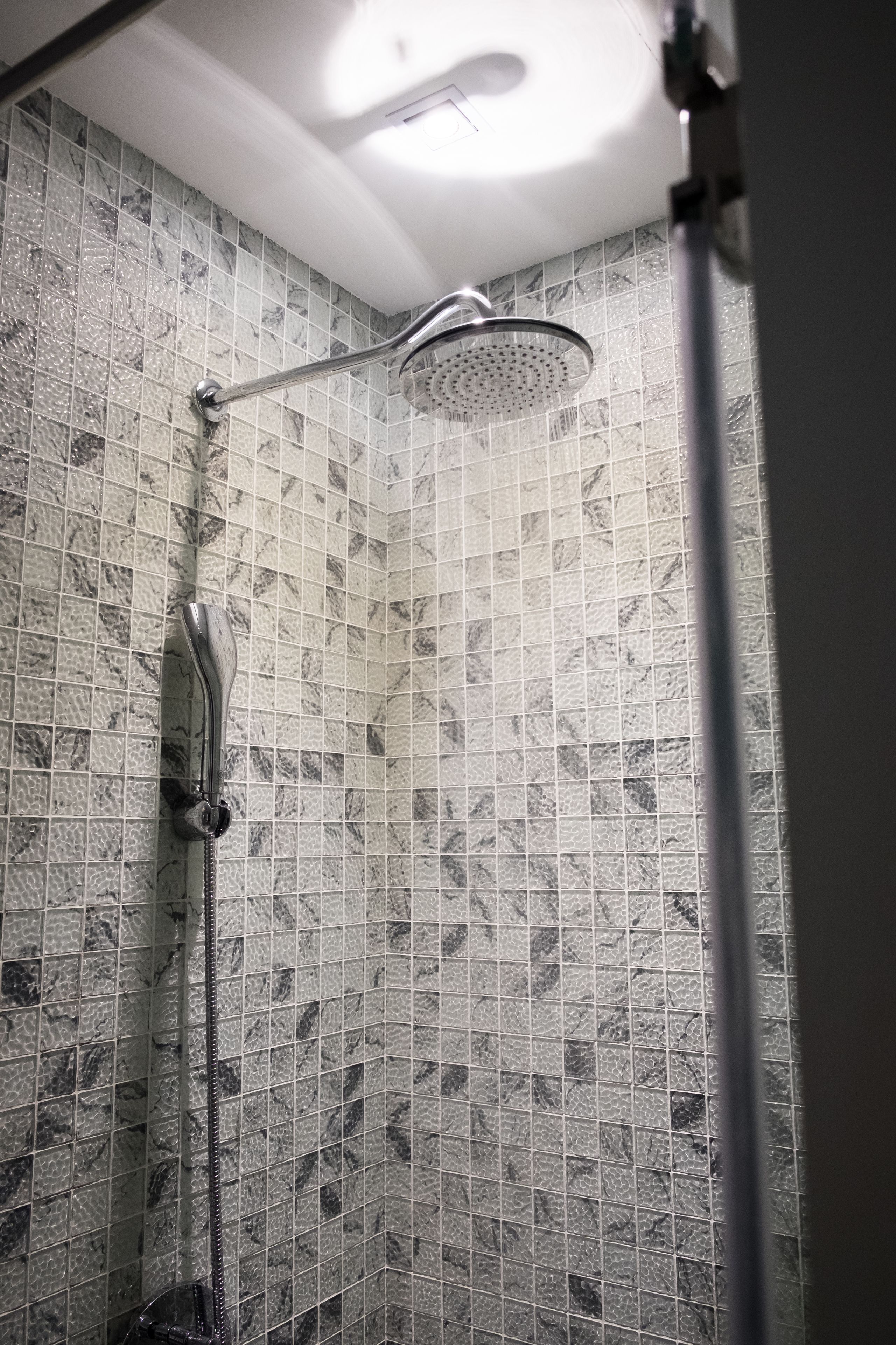 A shower with a handheld showerhead and a fixed overhead showerhead, tiled walls with a marble pattern, and a ceiling light fixture.