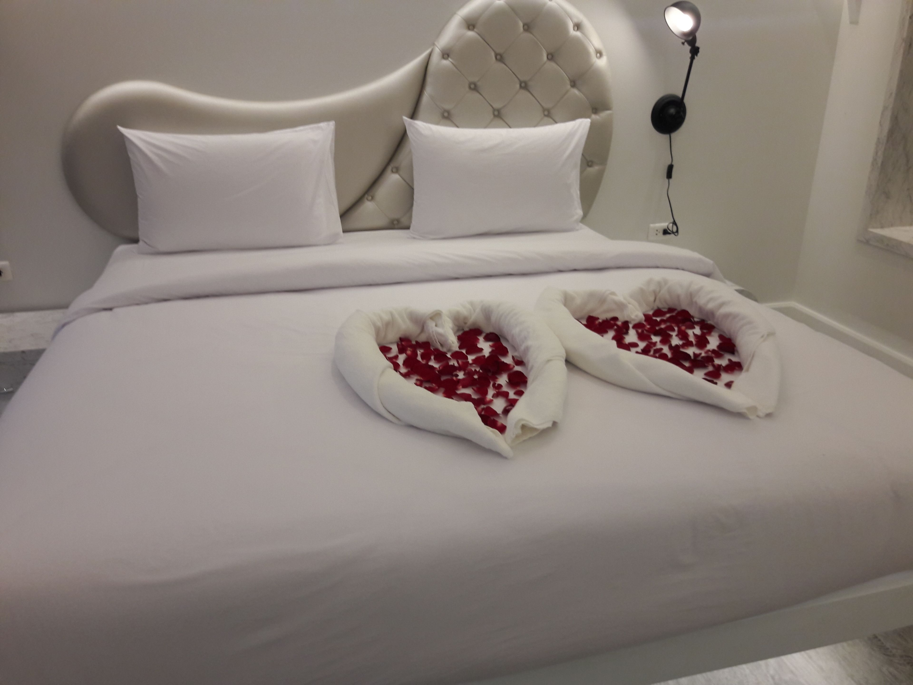 Una cama con sábanas blancas, un cabecero capitonado y dos arreglos de pétalos de rosa en forma de corazón sobre la cama.