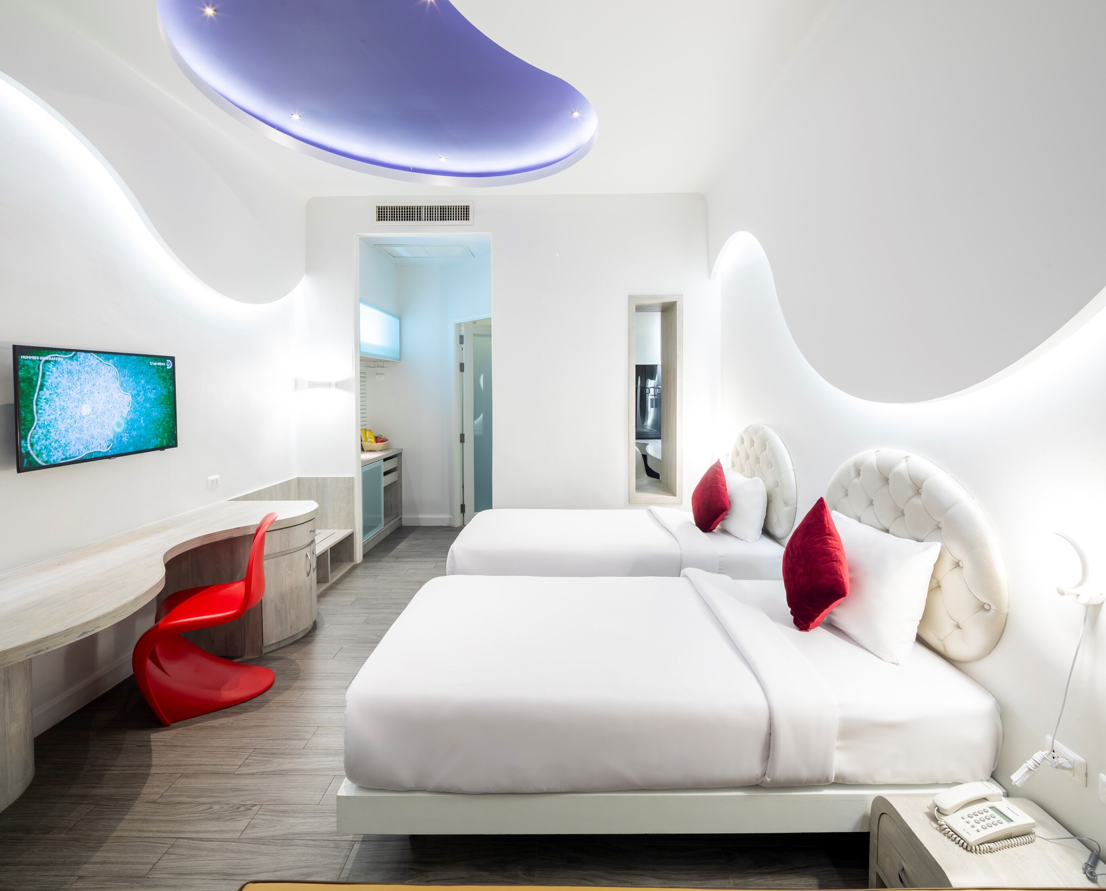 A modern hotel room with a flat-screen TV, a desk, two beds with red pillows, and a unique ceiling design.