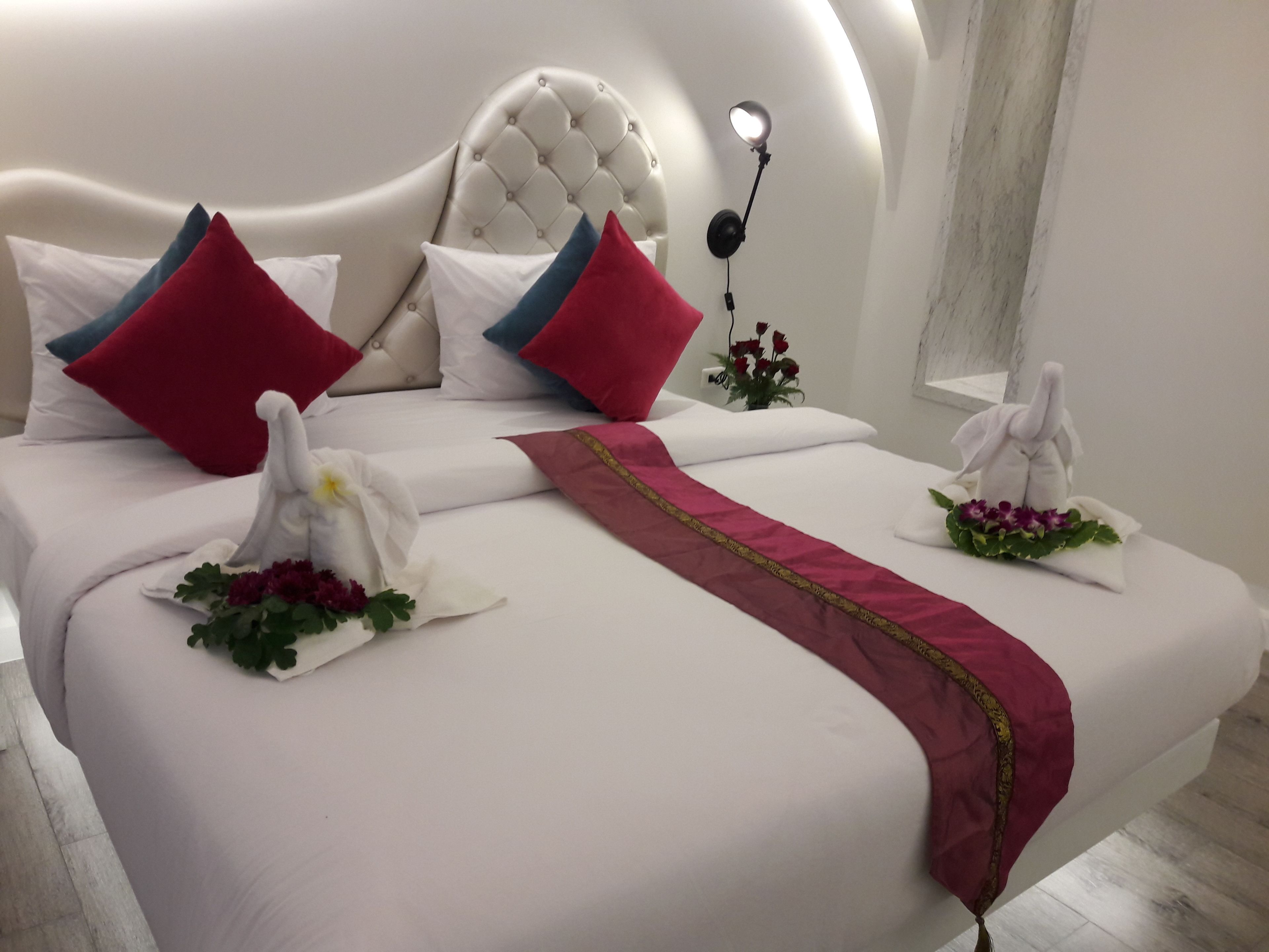 Un lit bien fait, avec du linge de lit blanc, des coussins rouges et bleus, et des serviettes décoratives ornées de motifs floraux.