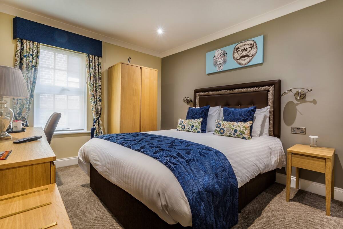 Une chambre à coucher avec un grand lit, une armoire en bois, un bureau avec une lampe et une fenêtre avec des rideaux.