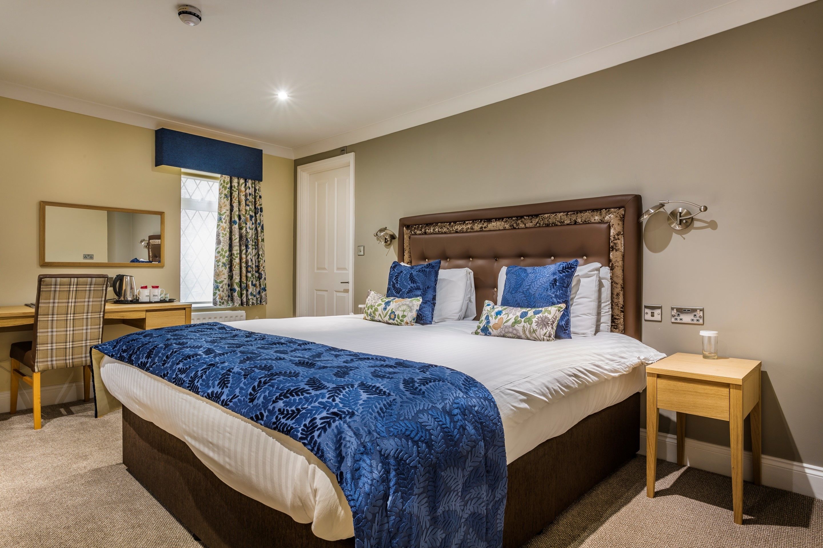 A neatly arranged bedroom with a large bed, a bedside table, a chair, a desk, and a mirror.