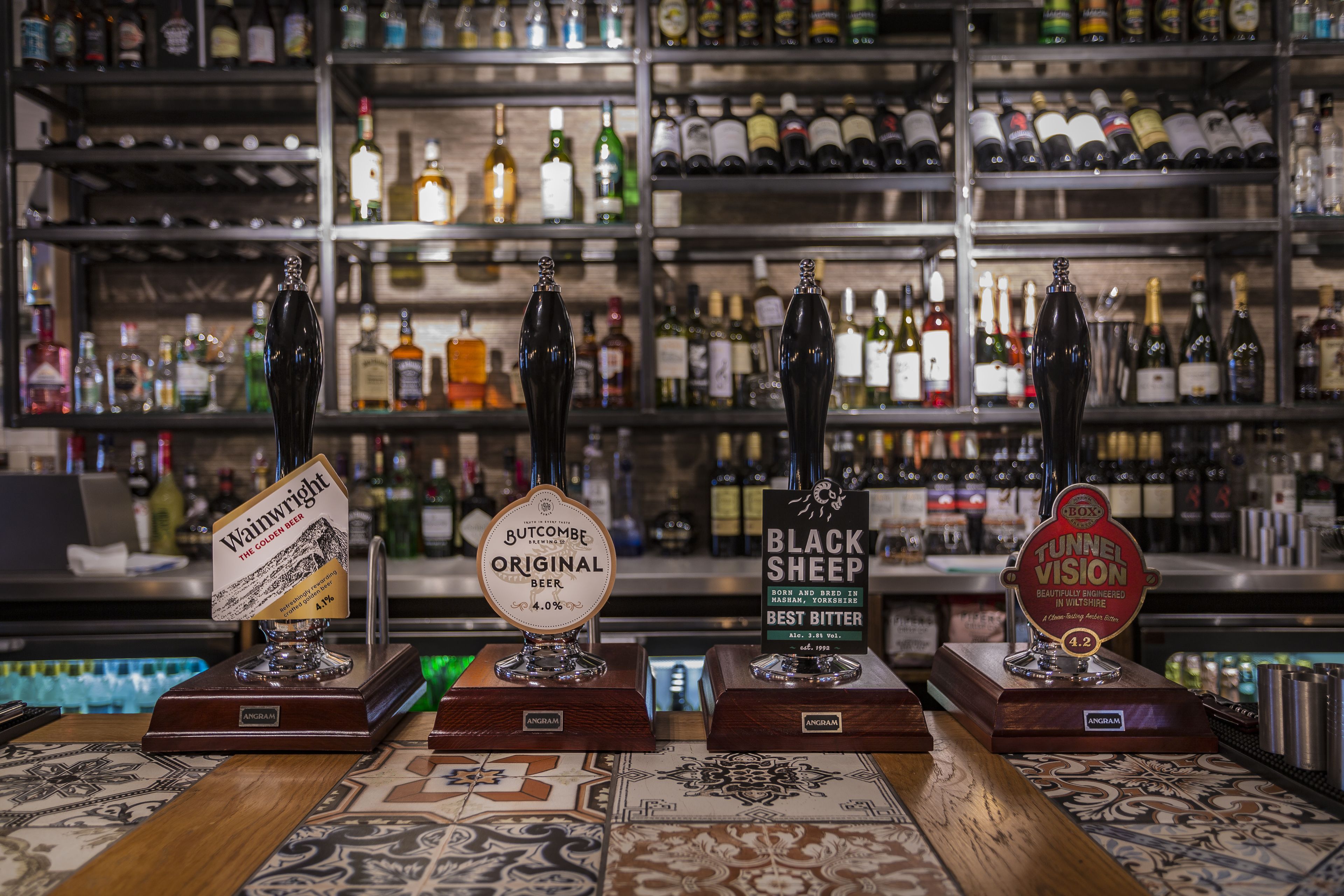 Cuatro grifos de cerveza con etiquetas en una barra, rodeados de estantes llenos de botellas diversas.