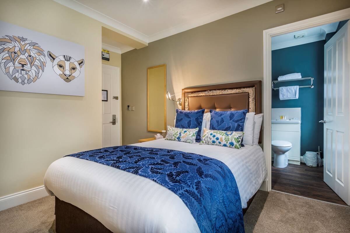Une chambre à coucher avec un grand lit, une tête de lit, une table de chevet et une salle de bain visible à travers une porte ouverte.