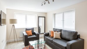 A modern living room with a glass coffee table, two black sofas with orange and beige cushions, a floor lamp, and large windows with blinds.