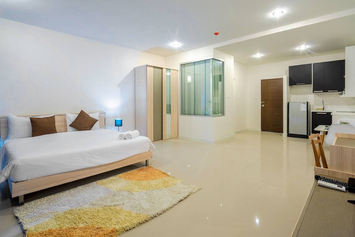 A modern bedroom with a large bed, a small desk, and a kitchenette.