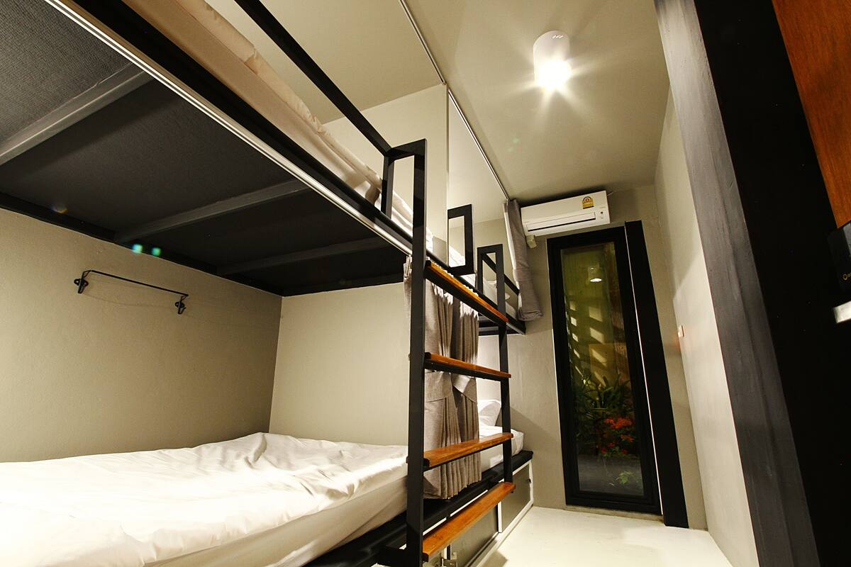 A compact hotel room with a bunk bed, a small window with curtains, and a built-in air conditioning unit.