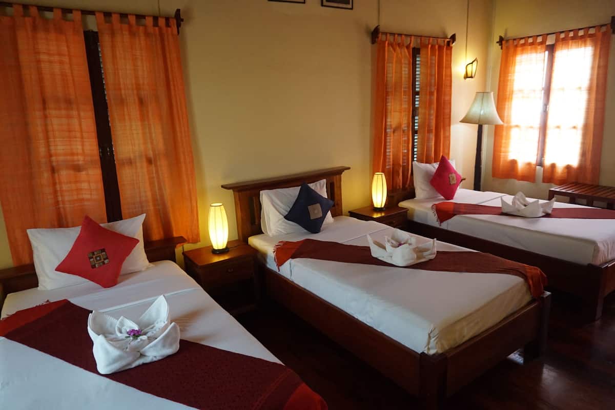 Habitación de hotel con tres camas individuales, cada una con almohada roja y blanca, cabecera de madera y mesita de noche con lámpara.