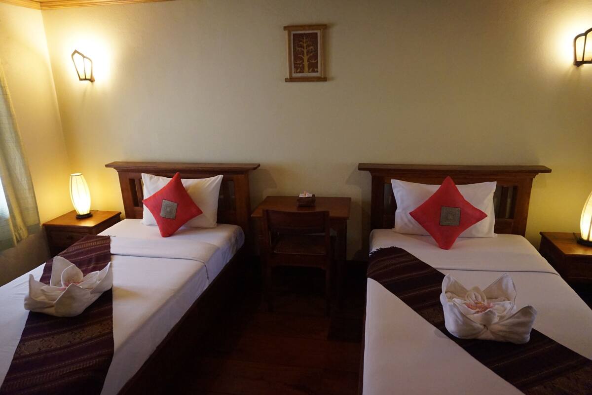Habitación de hotel con dos camas individuales, cada una con cabecero de madera, una mesita pequeña de madera y una mesita de noche con lámpara.