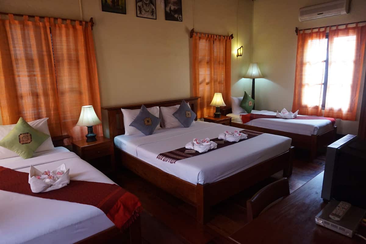 Habitación de hotel con tres camas, cada una con almohada y lámpara de noche. Hay un televisor sobre un mueble bajo y una ventana con cortinas.
