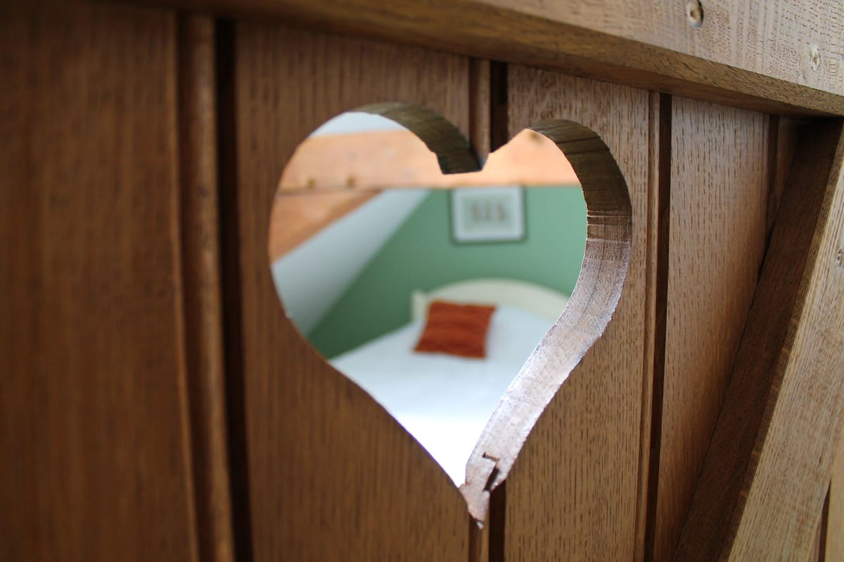 A heart-shaped window in a wooden structure, revealing a bed with a white sheet and a red pillow.