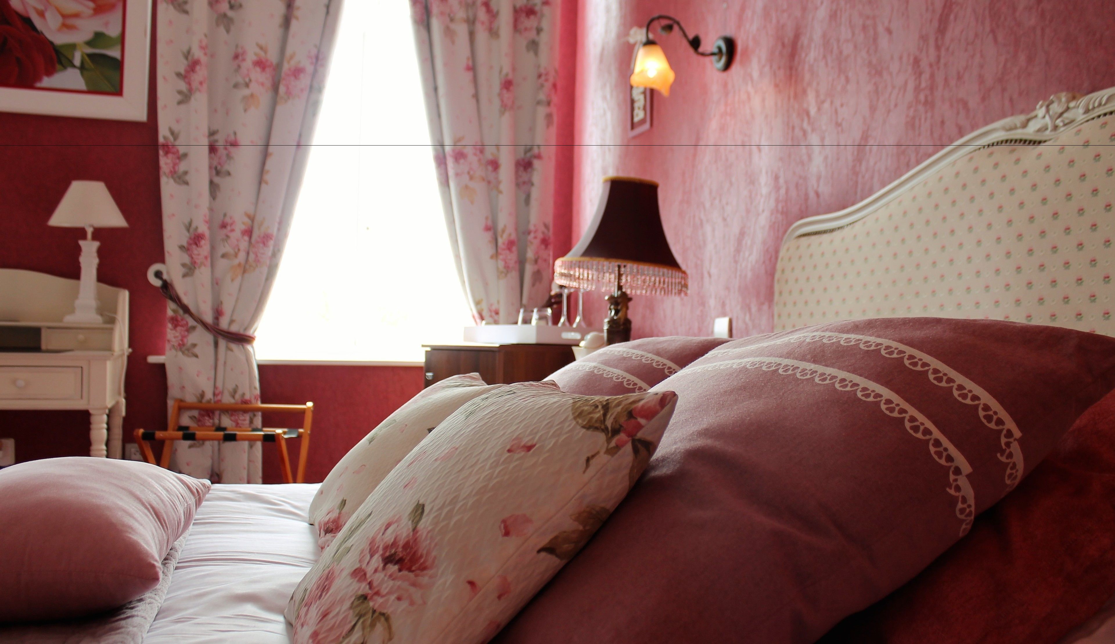 Ein Schlafzimmer mit blumigem Bettbezug, einer Nachttischlampe und einem Fenster mit Vorhängen.