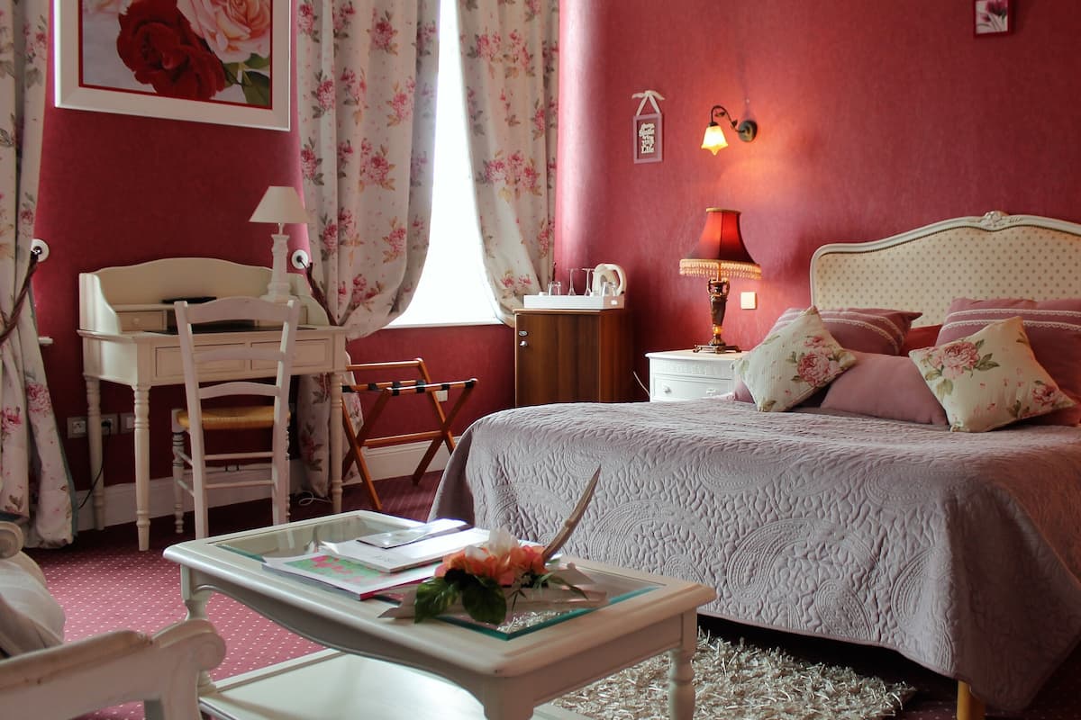 A bedroom with a bed, a desk, a chair, a lamp, and a window with floral curtains.