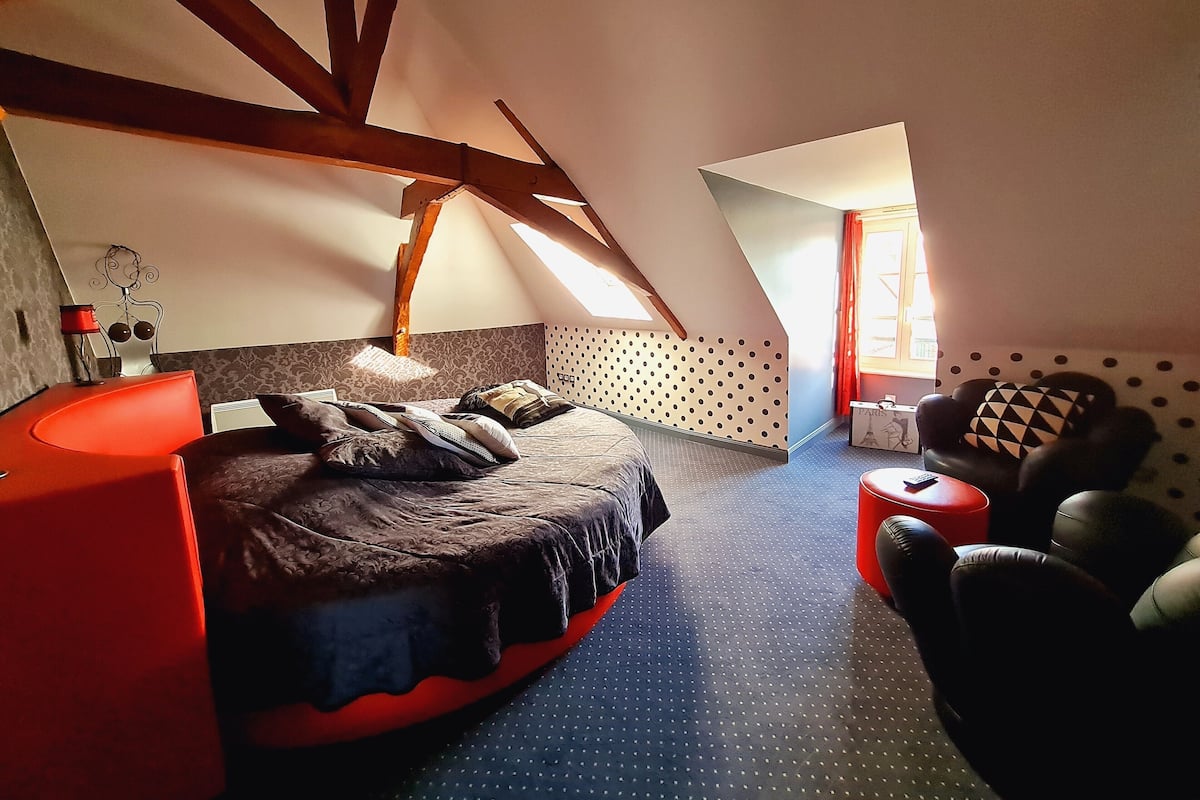 A bedroom with a sloped ceiling, a bed with multiple pillows, a red chair, and a small table.