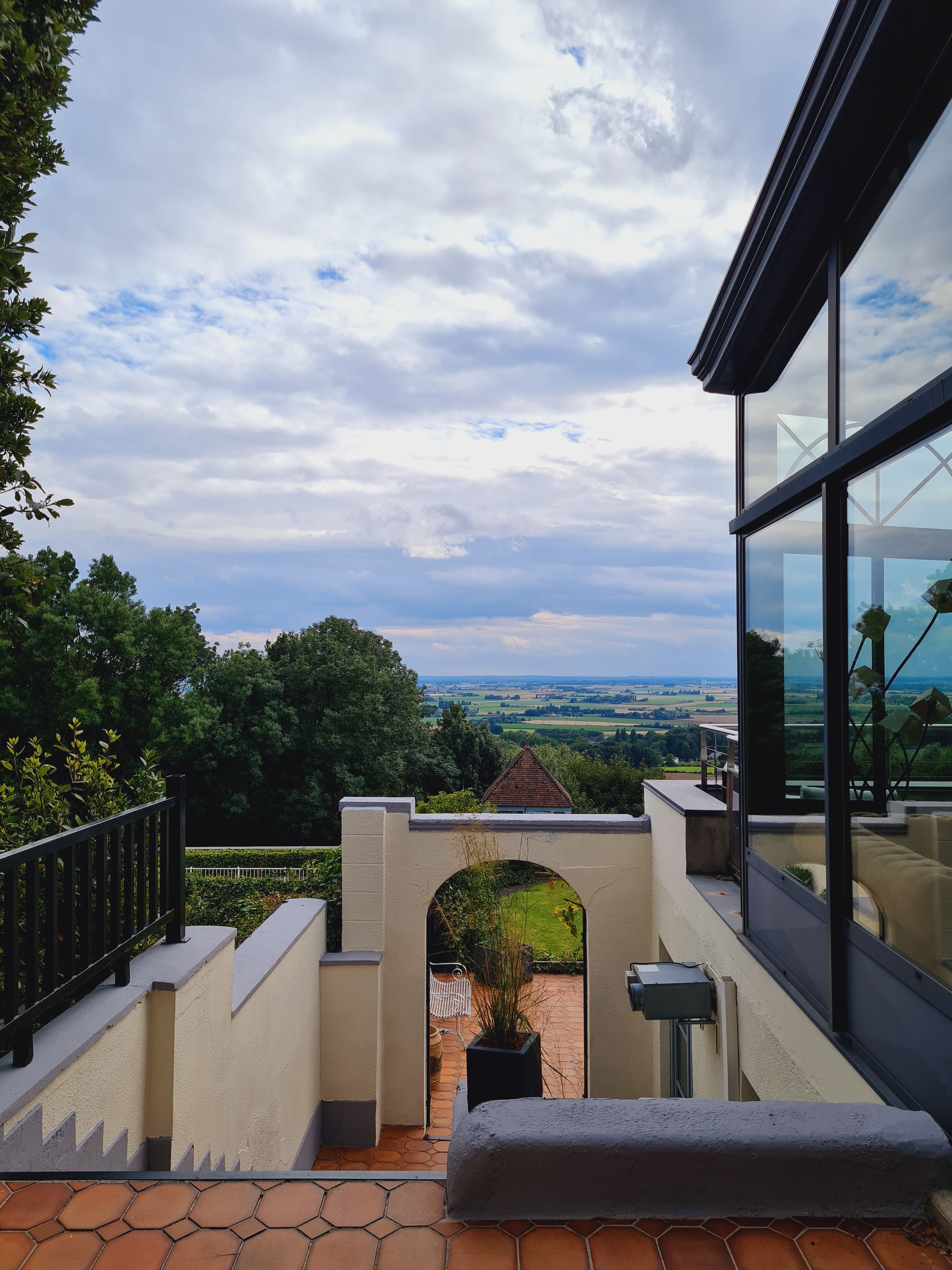 Un bâtiment moderne doté d’une façade en verre, offrant une vue sur la campagne, et d’un espace extérieur pavé agrémenté d’une rambarde.