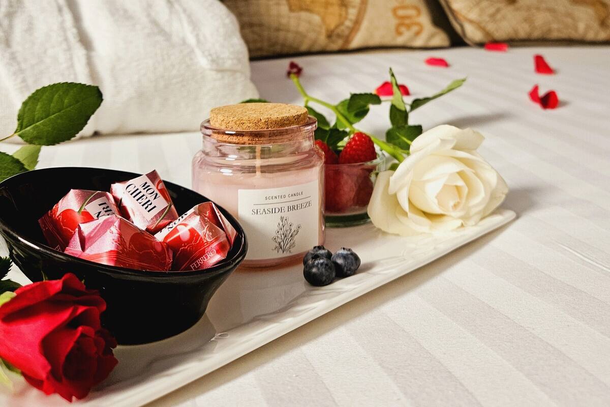 A neatly arranged tray with a jar of scented candle, a bowl of chocolates, a white rose, and scattered rose petals.