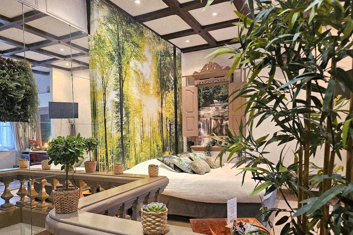 A hotel room with a large forest mural, a bed with multiple pillows, a small table with a menu, and a balcony with plants.