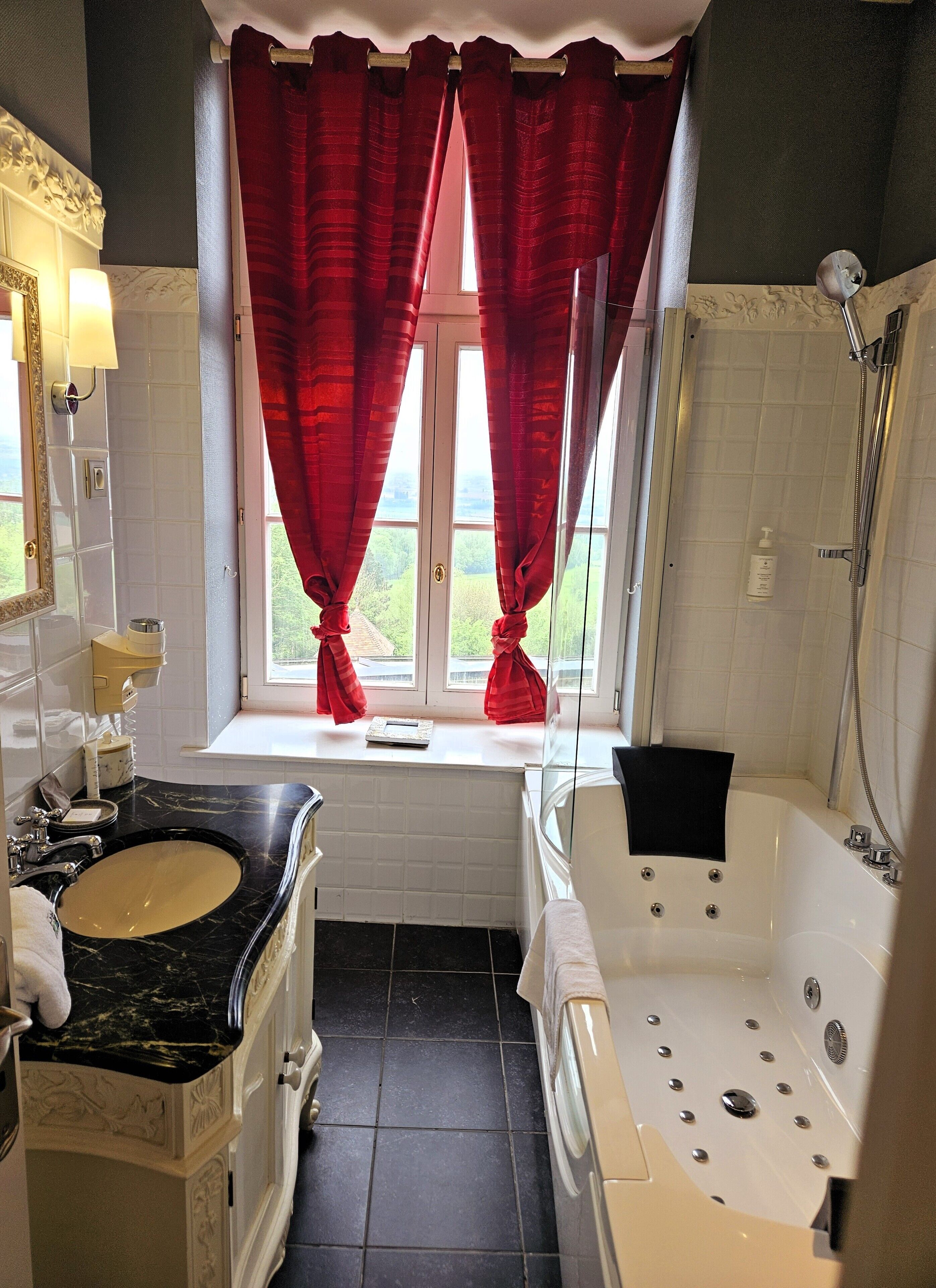 A bathroom with a marble sink, a bathtub, and a window with red curtains.