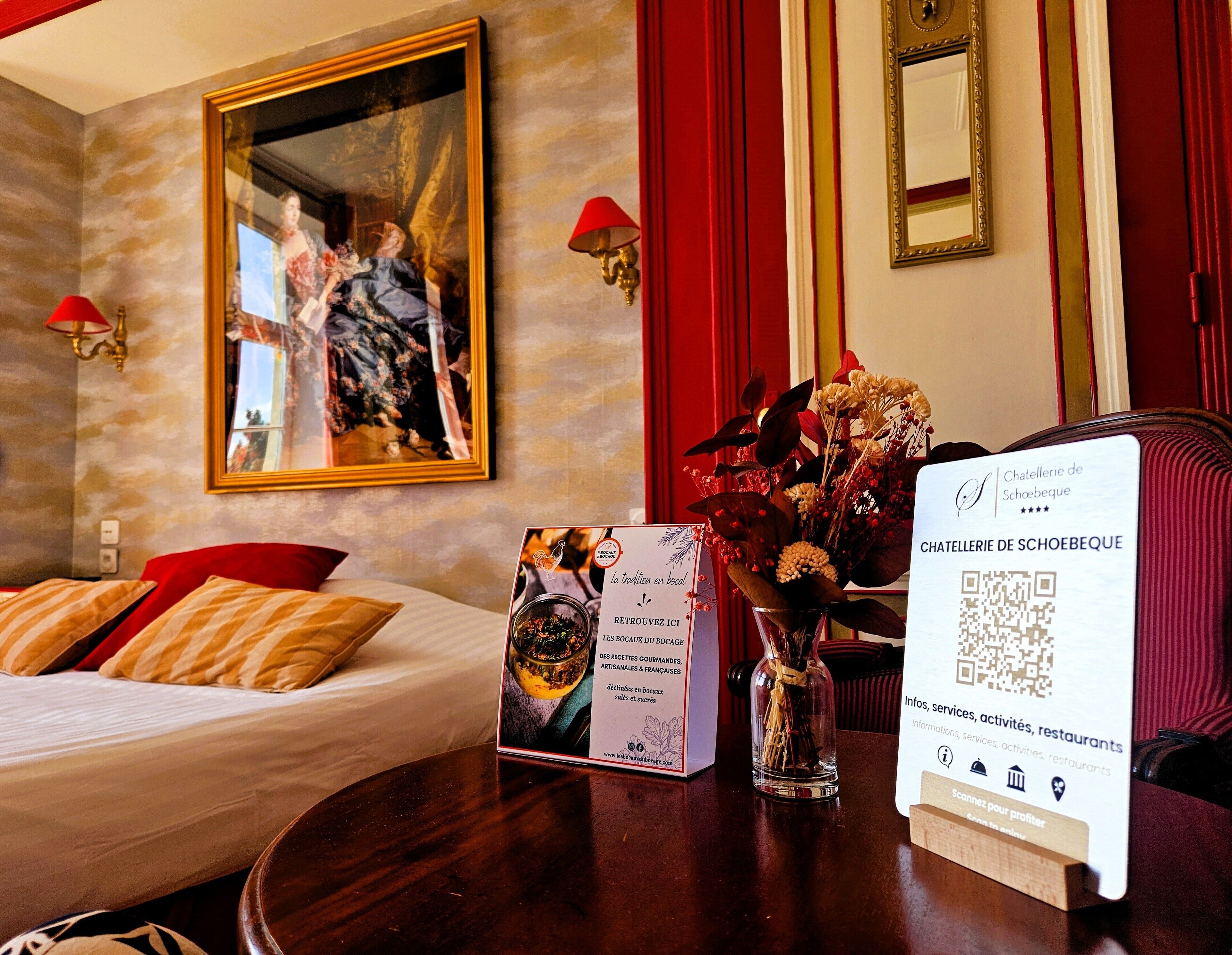 Habitación de hotel con una cama, un cuadro enmarcado, un florero con flores, un folleto y un cartel con un código QR.