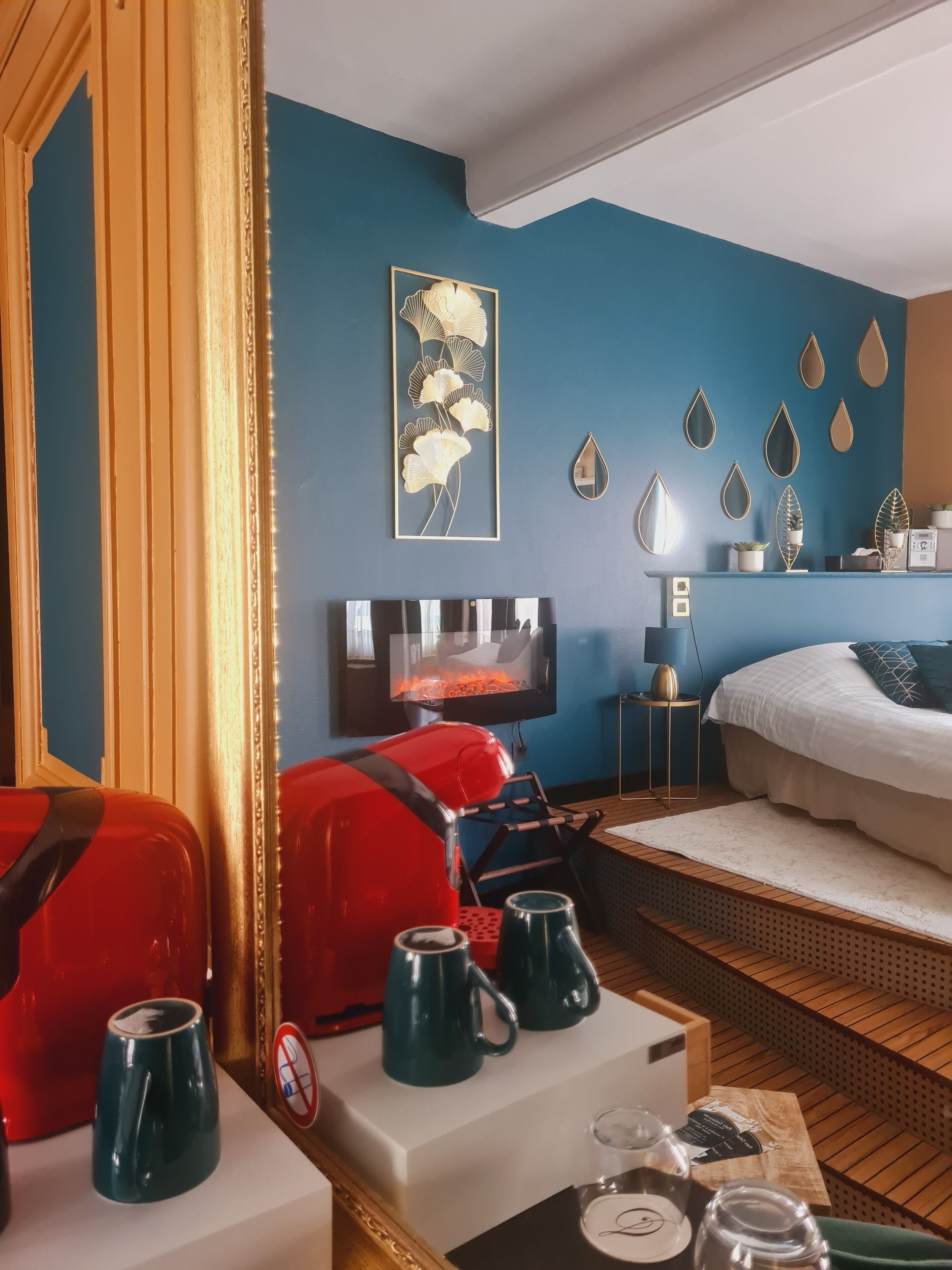 A hotel room with a bed, a TV, a coffee table with mugs, a red chair, and a wall with decorative items.
