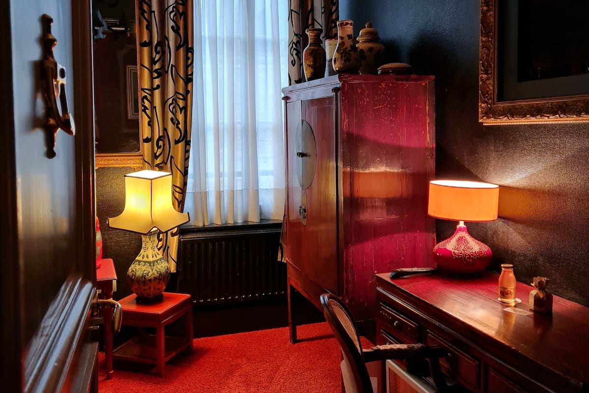 A room with a red carpet, a wooden desk with a lamp, a chair, a cabinet, and a window with curtains.