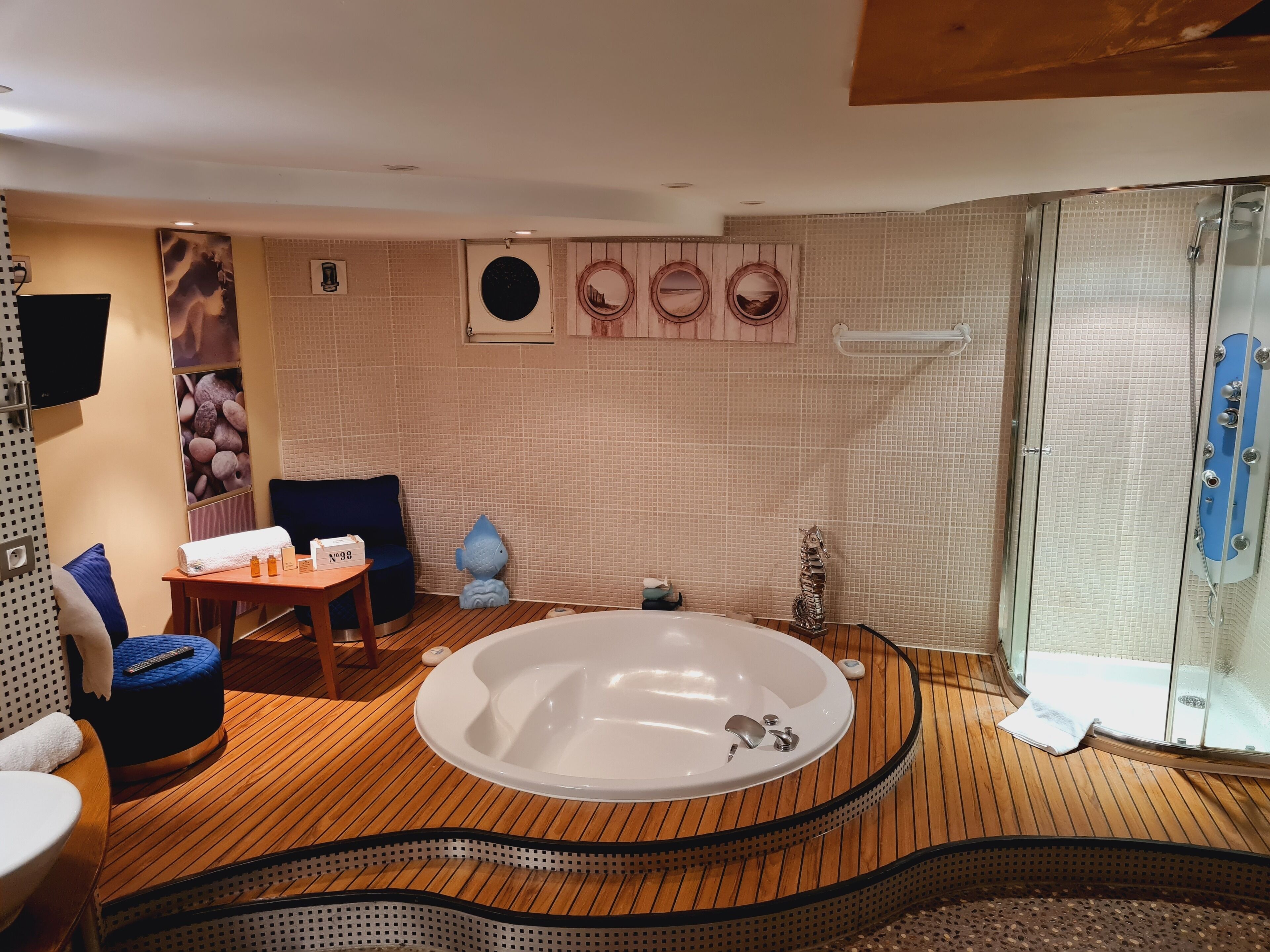 Un área de spa con piso de madera, una tina de hidromasaje ovalada grande, una ducha con cerramiento de vidrio y una zona de estar con mesa y sillas.