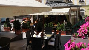 An outdoor seating area with tables and chairs, a covered patio, and a building with a sign reading "HOTEL ROZE". - Hotel Malta (Karlovy Vary)